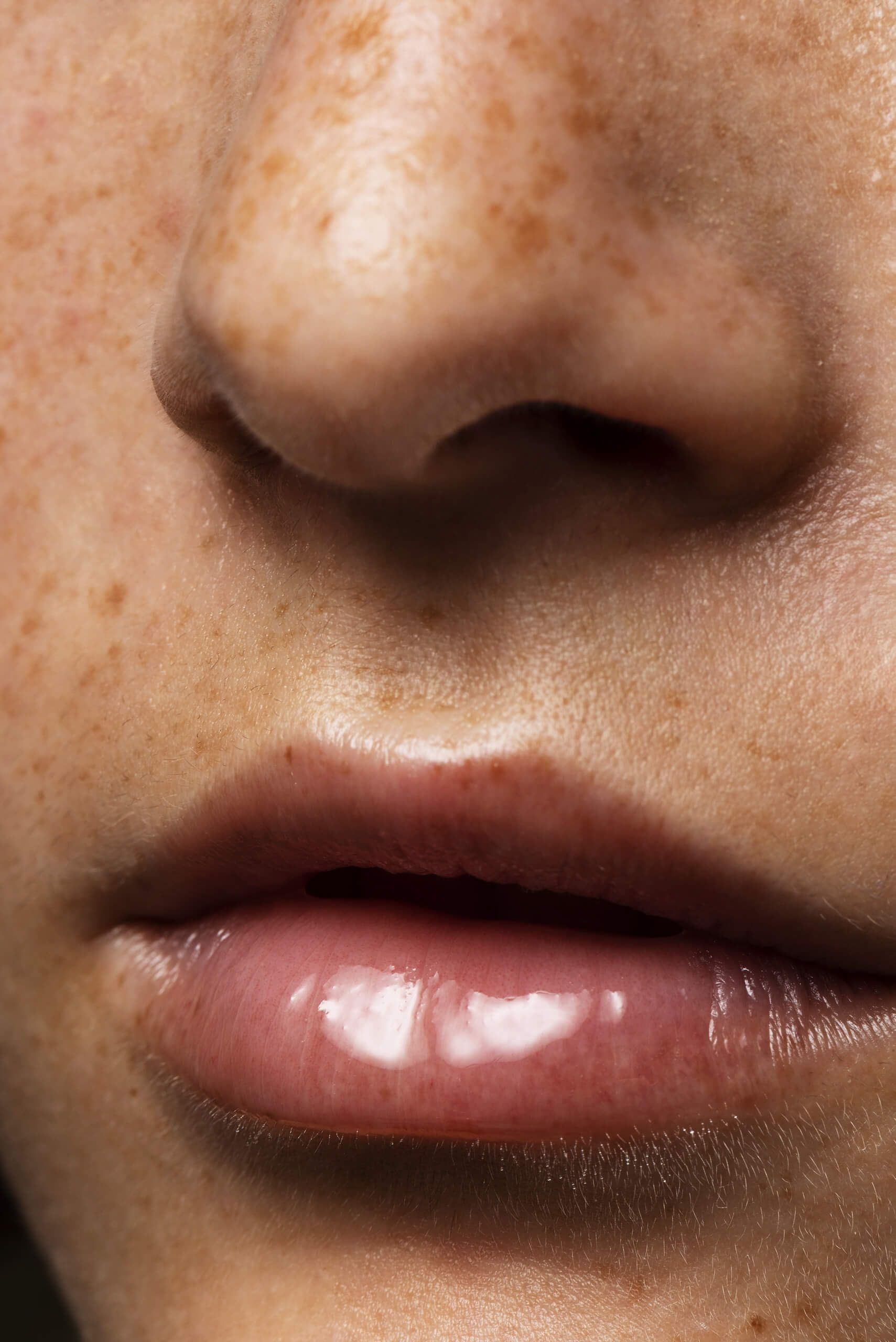 Close-up di labbra idratate con effetto lucido, leggermente socchiuse, e pelle del viso con lentiggini visibili