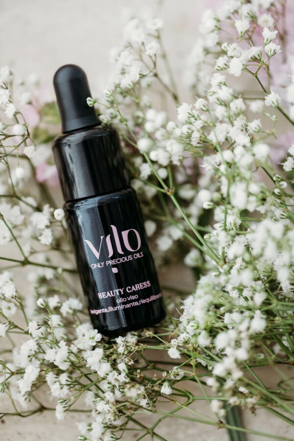 Foto di un boccetto di olio viso beauty caress di vilo cosmetics in mezzo a rametti con fiorellini piccoli bianchi