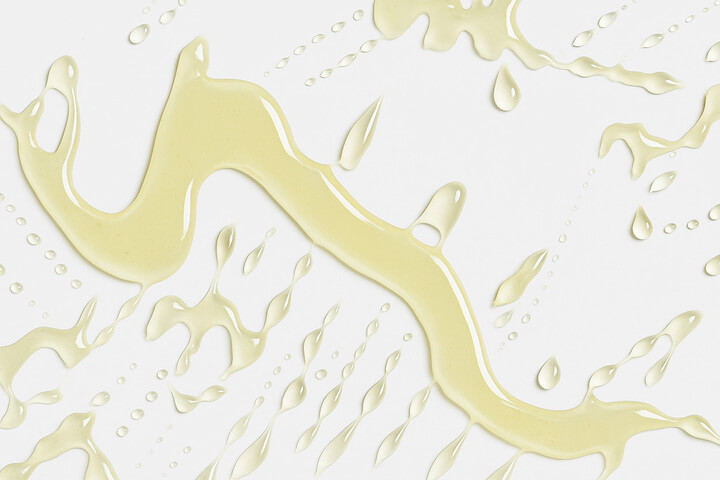 Texture oleosa della vitamina E contenuta nel balsamo labbra Vilo Lip Oil, con gocce e striature su sfondo bianco che evocano idratazione e nutrimento